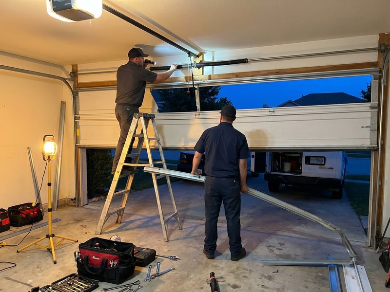 Two technicians in dark uniforms working on a partly raised door at dusk, one of them on a ladder