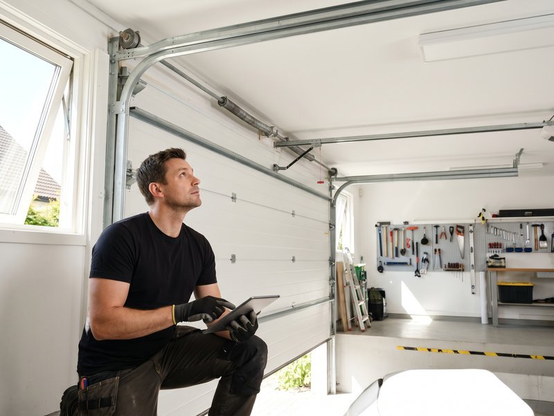 Technician kneeling in a garage and checking a white sectional door with a tablet