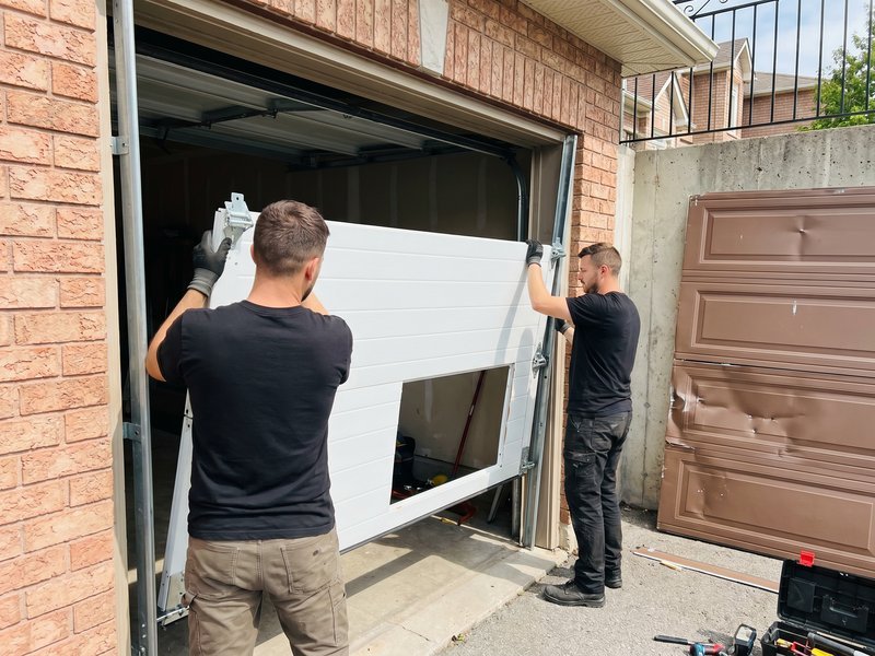 Two technicians lifting a new panel into a brick-framed garage opening